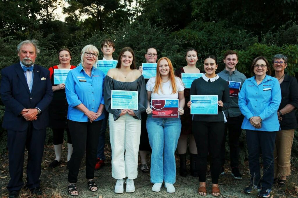 A group of students are holding up their awards from the education support funds, surrounded by two bank representatives and members of the community foundation. The light is dark against a bushy background of trees.