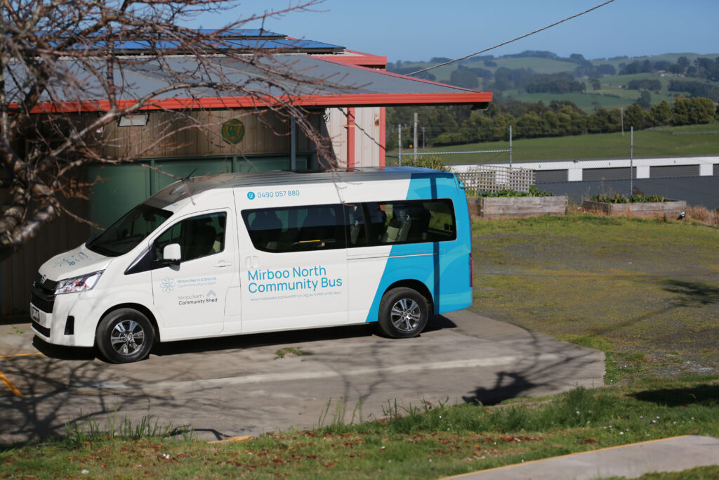 Image of white van, parked under cover on sunny day. Blue panel at read wheel with words in blue written across side door that reads, "Mirboo North Community Bus".