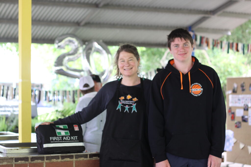 a woman and teenage boy stand next to a black first aid kit, the woman has one hand resting on the bag. they are smiling.