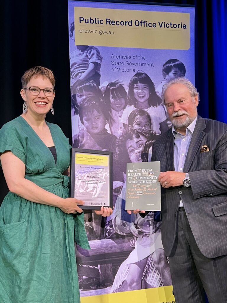 Two individuals, a woman on the left and a man on the right, are smiling and holding a commendation certificate and a book titled "FROM RURAL HEALTH TO COMMUNITY STRENGTHENING: The History of the Mirboo North & District Community Foundation". They are standing in front of a digital screen displaying a message of congratulations and thanks for contributions to the state's history, likely at the Victorian Community History Awards ceremony. The woman is wearing a green dress and the man is in a smart suit with a patterned tie. The mood is celebratory and proud.