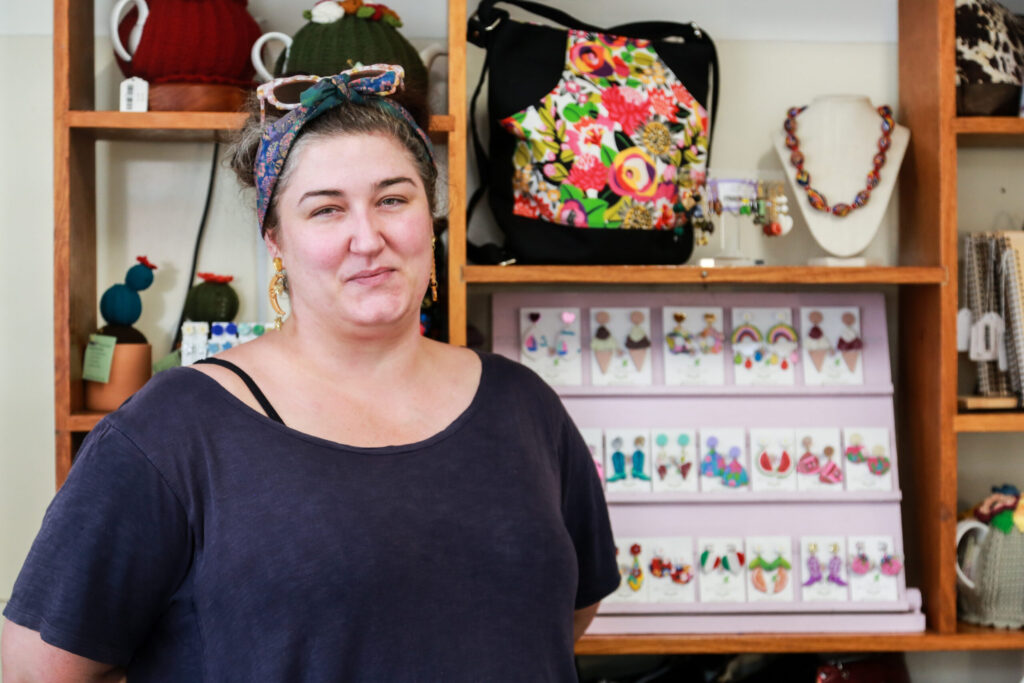 artist of workshop, Kate Billingsley stands in front of her polymer clay jewelry in a headscarf and dark grey tshirt.