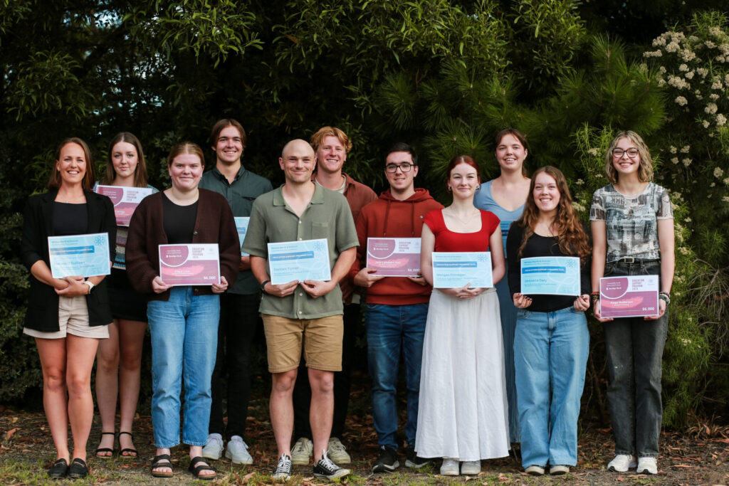 A group of students in order of tallest at the back and shortest at the front are arranged holding certificates of grants awarded, in front of a green bushy background