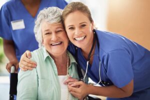 Smiling elderly woman sitting in a wheelchair embraces a cheerful female nurse in blue scrubs and a stethoscope, both looking directly at the camera. Another healthcare worker stands slightly out of focus in the background, also wearing blue scrubs.