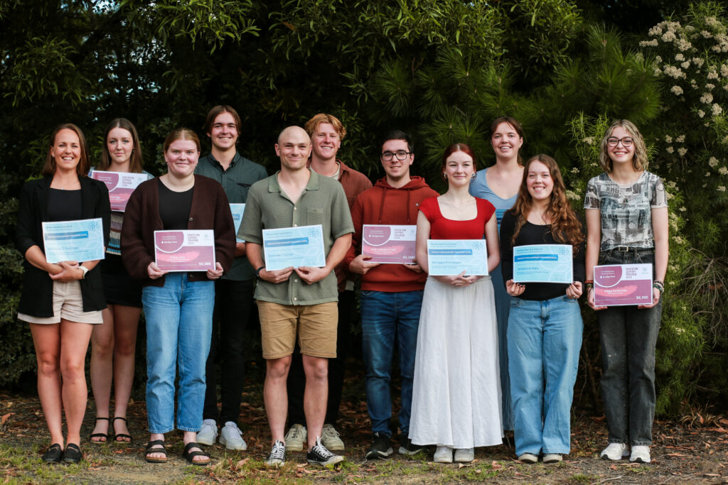 A group of award recipients stand in a group for a photo, holind their certificates in order of height in front of a green bushy tree all smiling.