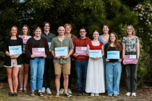 A group of award recipients stand in a group for a photo, holind their certificates in order of height in front of a green bushy tree all smiling.