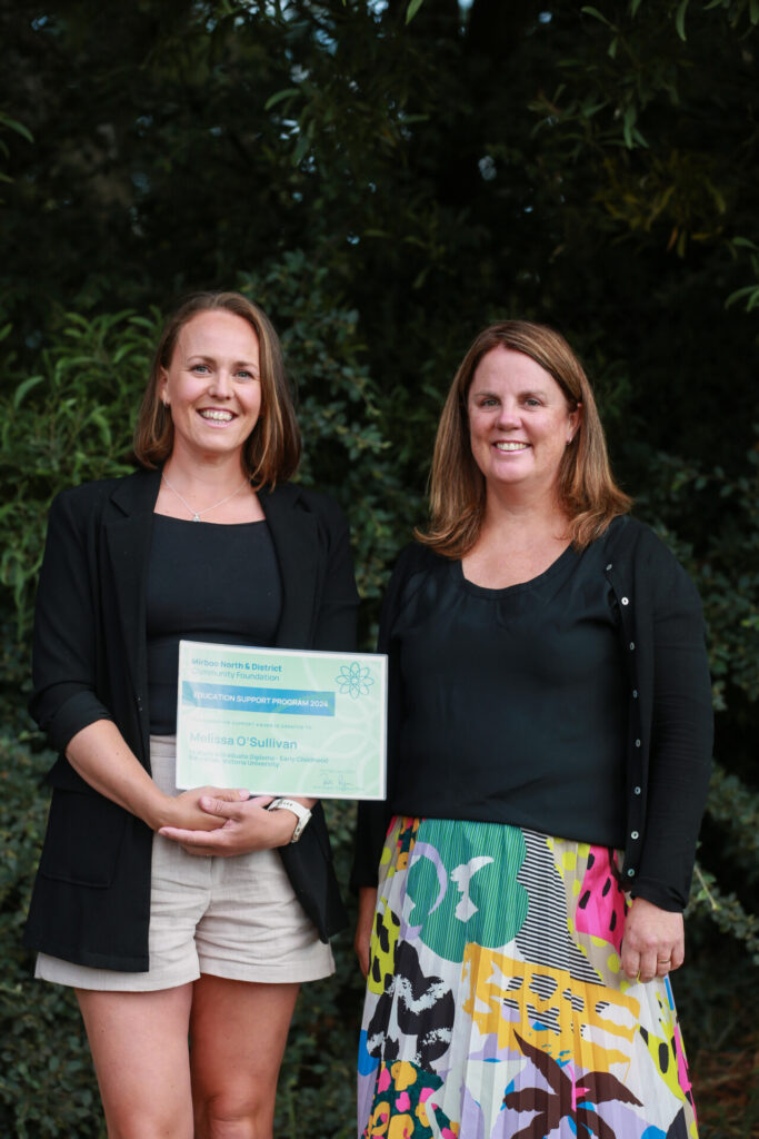 two women stand beside each other, the woman on the right holds a certificate and the woman on the right wears a colourful skirt
