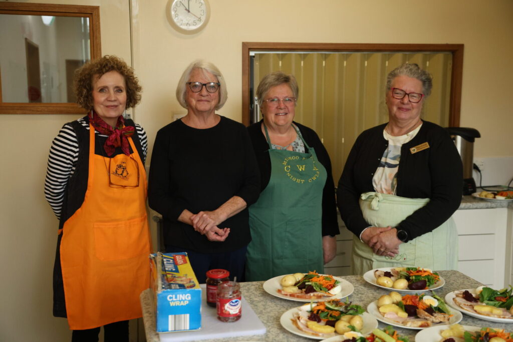Four women stand inside a kitchen wearing aprons, posing for a photo with their hands in front of their bodies with plates of food spread out in front of them.
