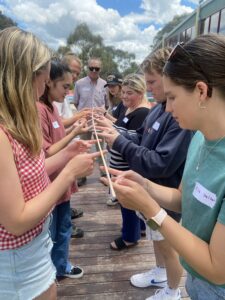 two lines of young adults stand facing each other each holding out their index fingers to hold up a long piece of string symbolising team work while team leader stands down the centre observing.