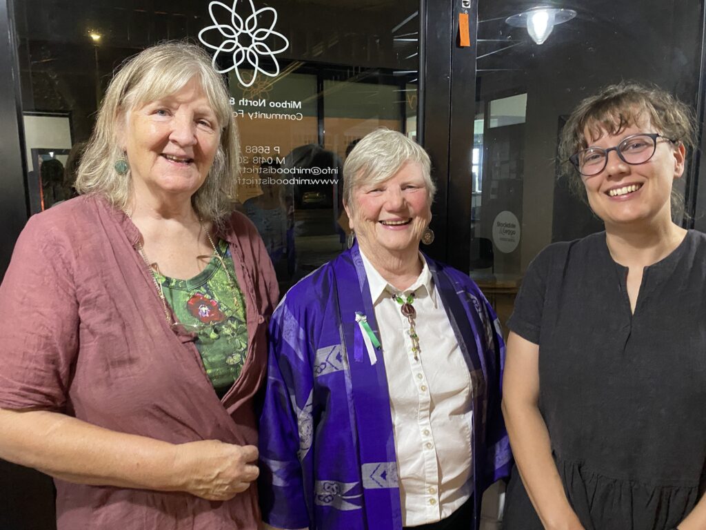 Three woman stand smiling facing the camera indoors with a glass window behind them.