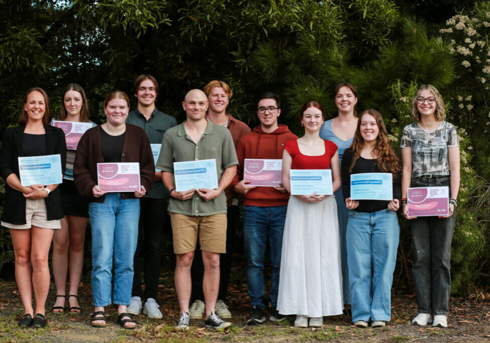 A group of award recipients stand in a group for a photo, holind their certificates in order of height in front of a green bushy tree all smiling.