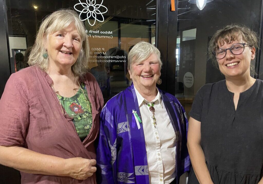 Three woman stand smiling facing the camera indoors with a glass window behind them.