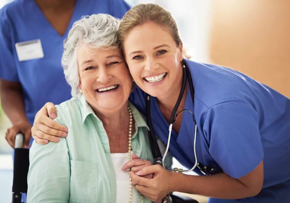 Smiling elderly woman sitting in a wheelchair embraces a cheerful female nurse in blue scrubs and a stethoscope, both looking directly at the camera. Another healthcare worker stands slightly out of focus in the background, also wearing blue scrubs.
