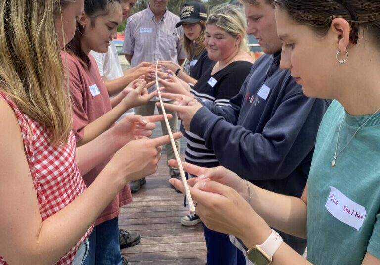 two lines of young adults stand facing each other each holding out their index fingers to hold up a long piece of string symbolising team work while team leader stands down the centre observing.
